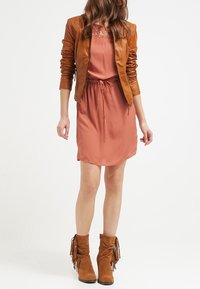 Chaqueta de cuero marrón sobre un vestido color coral con detalles de encaje y cintura con cordón, combinado con botas de tobillo marrones con flecos.