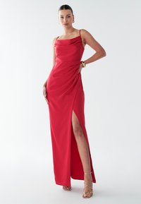 Červené večerné šaty s plášťovým výstrihom, tenkými ramienkami a rozparkom až na stehno. Vyrobené z hladkej látky s nariaseným detailom na boku.
