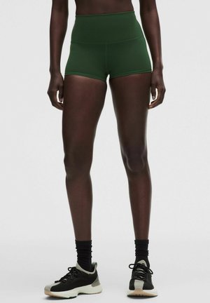 Persona con pantalones cortos deportivos de cintura alta de color verde oscuro, calcetines negros hasta el tobillo y zapatillas de correr en blanco y negro, de pie sobre un fondo liso.