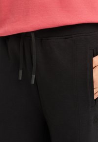 Svarta sweats byxor i mjukt tyg, med elastisk midja och snodd samt sidofickor. Texturerad logotypdetalj vid fickan.