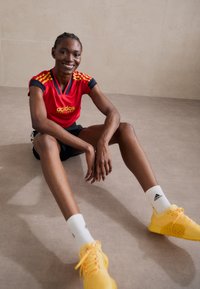 Sarkans īsajām piedurknēm Adidas sporta krekls ar dzeltenām akcentiem, apvienots ar melniem šortiem. Modelis valkā dzeltenas sporta kurpes un baltas zeķes.