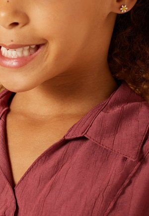 Chemise texturée bourgogne avec un col, arborant une petite boucle d'oreille florale. Gros plan sur la partie inférieure du visage et le cou d'un enfant.