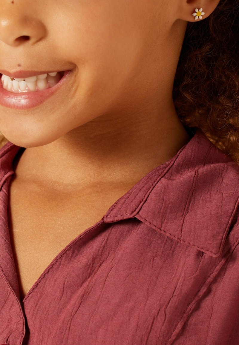 Bordeauxkleurig gestructureerd shirt met een kraag, voorzien van een kleine bloemenoorbel. Close-up van de onderkant van het gezicht en de nek van een kind.