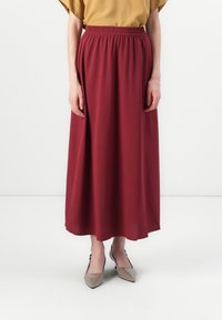 Jupe longue bordeaux avec une taille élastique et des poches latérales, en tissu doux, associée à des escarpins beiges à bouts pointus.