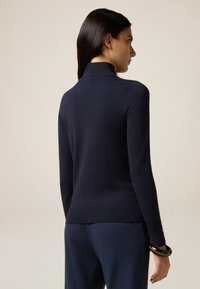Marineblauwe, aansluitende trui met hoge kraag, geribde manchetten en zoom. Gladde textuur; gecombineerd met donkerblauwe broeken en een armband om de pols.