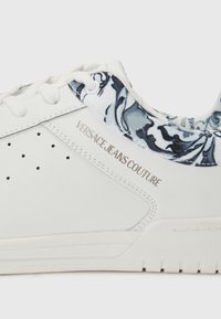 Hvite sneakers med en glatt lærbekledning og en blå marmorert tekstilkrage, med sølvfarget merkevare på siden og perforerte detaljer.