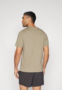 Tricou scurt de culoare maronie deschis, cu guler circular, textură netedă și tiv drept, purtat cu pantaloni scurți negri, văzut din spate.