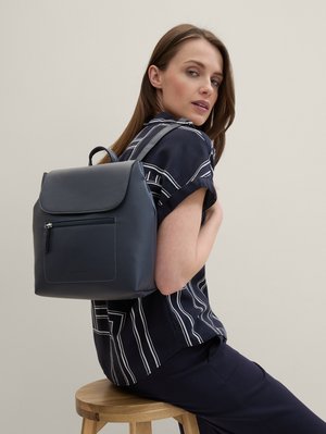 Frau, die auf einem Holzstuhl sitzt, trägt ein navy-blau gestreiftes Hemd und einen navy-blauen Leder-Rucksack über einer Schulter vor einem beigen Hintergrund.
