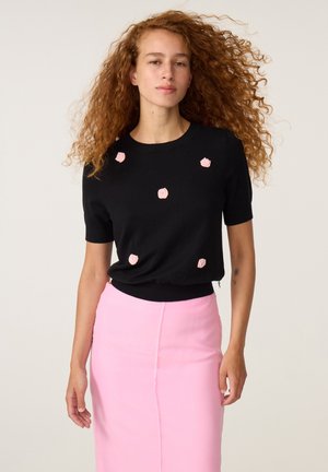 Vrouw met lang krullend haar die een zwarte top met korte mouwen draagt met roze bloemendetails en een roze rok met hoge taille.
