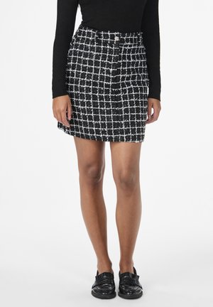 Jupe mini en tweed à carreaux noir et blanc avec une finition texturée. Elle présente une taille ajustée et un ourlet léger. Portée avec des chaussures noires.