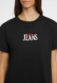 T-shirt in cotone nero con testo "guess JEANS" in bianco e rosso, collo tondo e maniche corte. Design semplice, texture liscia.