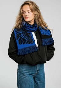 Niebiesko-czarny szalik z motywem paproci, z frędzlami, noszony na czarnej koszuli. Casualowy design o miękkiej fakturze.