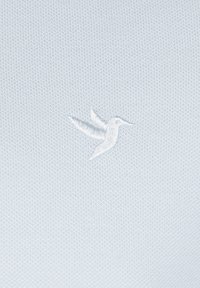 Himmelblau gestrickter Stoff mit einem kleinen, weißen gestickten Kolibri-Logo, das versetzt positioniert ist. Schlichte und strukturierte Oberfläche.