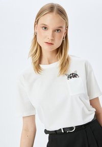 Blonde Frau mit silbernen Creolen, die ein weißes T-Shirt und schwarze Hosen trägt, während eine Katze aus der Brusttasche des Shirts hervorschaut.