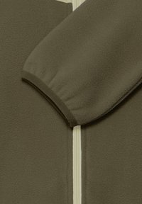 Gros plan de la manche d'une veste polaire vert olive avec un poignet élastique et une fermeture éclair blanche visible à l'avant.