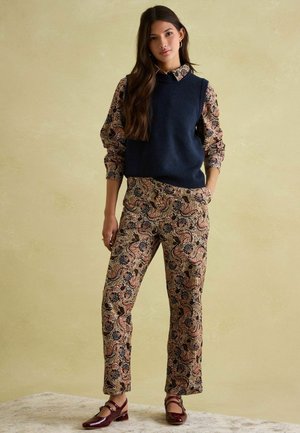 Navy knitted vest over a patterned blouse and matching trousers in earthy tones. Shoes are maroon with a low block heel. Background is light.