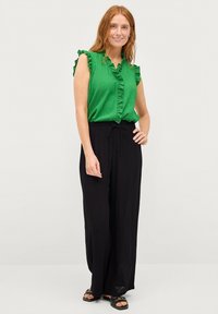 Groene mouwloze top met ruches bij de hals en zwarte wijde broek. Gladstoffen textuur; de outfit heeft een ontspannen pasvorm, een gestrikt middel en sandalen.