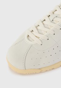 White leather sneaker with perforated detail, cream laces, and a textured beige rubber sole. Features clean stitching and minimal design.