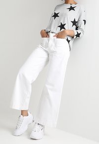 Maglione grigio con motivi a stella nera, abbinato a pantaloni bianchi a gamba larga e sneakers bianche, con una vestibilità rilassata e un design minimalista.