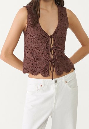 Bruine mouwloze gehaakte top met cirkelvormige patronen en drie knoopsluitingen aan de voorkant, gedragen met witte broek met hoge taille.