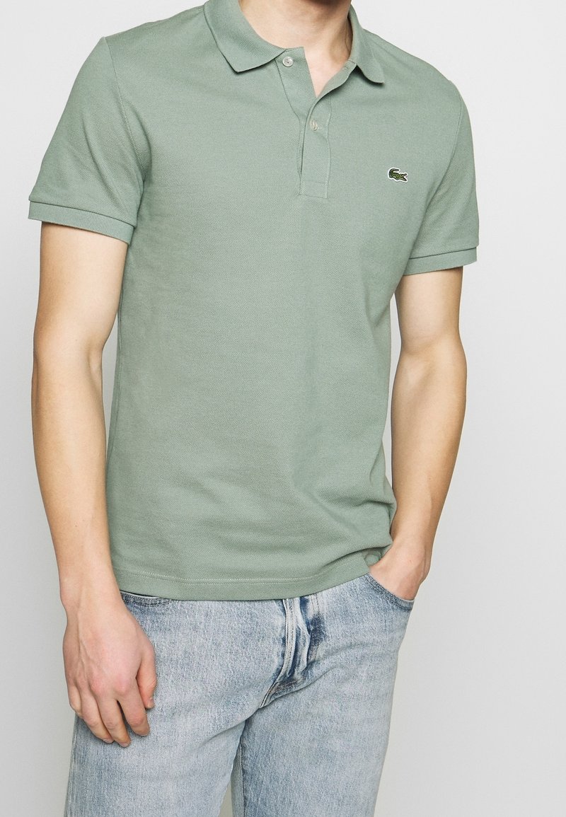 Homme portant un polo à manches courtes vert clair avec un petit logo de crocodile et un jean bleu clair, les mains dans les poches, sur un fond uni.