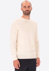 Uomo con barba e testa rasata che indossa un maglione a maniche lunghe color crema e pantaloni scuri, in piedi contro uno sfondo chiaro e uniforme.