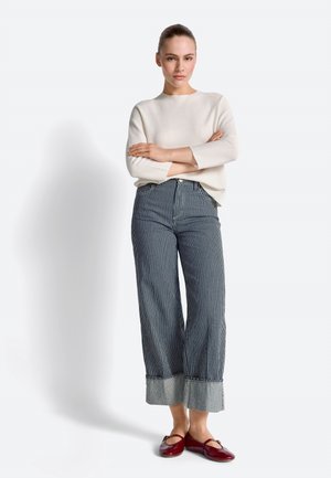 Woman standing with arms crossed wearing white sweater, high-waisted blue and white striped wide-leg pants, and red flats.