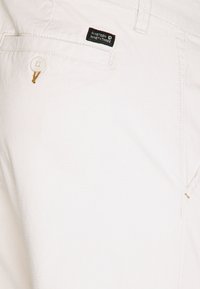 Pantalones de algodón blancos con un acabado texturizado, que presentan un bolsillo delantero adornado con un botón y una etiqueta de la marca que dice "Nineteen Ninety-Three".