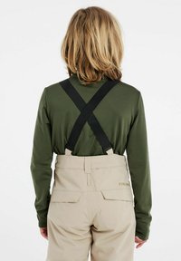 Beige Latzhose mit verstellbaren schwarzen Trägern, getragen über einem grünen Langarmshirt, mit Gesäßtaschen und einem schlichten, funktionalen Design.