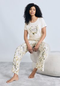 Σετ πυτζάμες ανοιχτό κρεμ με κοντομάνικη μπλούζα με εμπριμέ λουλούδια και το κείμενο "Πάντα ανθίζω" και παντελόνι με μοτίβο κίτρινα λουλούδια.