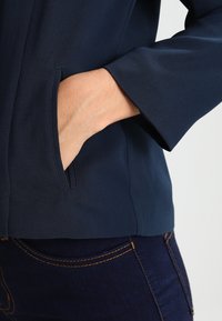 Marineblauwe jas met een gladde textuur en een discreet zijzakje; heeft een ontspannen kraag en licht uitlopende mouwen voor vrijer bewegen.