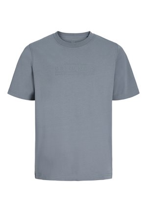 Ragazzo biondo in piedi e sorridente, indossa una maglietta grigia a maniche corte, pantaloni neri e sneakers bianche su sfondo bianco.