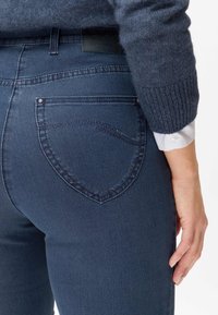 Jean de couleur bleu foncé avec une coupe ajustée, poches arrière visibles et une ceinture texturée. Le tissu semble lisse avec des accents de couture subtils.