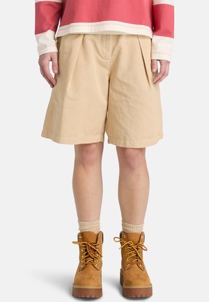 Beige plisserede shorts lavet af letvægtsstof, med en afslappet pasform, kombineret med brune snørestøvler og cremefarvede sokker.