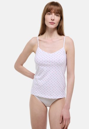 Jeune femme aux cheveux roux lisses, portant un caraco blanc orné de petits cœurs rouges et des sous-vêtements clairs, posant avec une main derrière le dos.