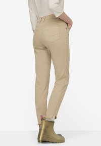 Béžové slim-fit kalhoty s zadními kapsami, které mají hladkou texturu a kontrastní šití, zkombinované s lesklými zlatými kotníkovými botami.