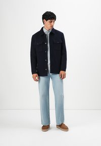 Mann in einer navyblauen Jacke mit Knöpfen über einem grauen Hemd, hellblauen Jeans und braunen Schuhen, der vor einem weißen Hintergrund steht.