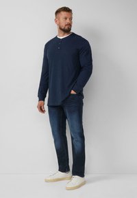 Marineblaue Langarm-Henley-Shirt mit einem weißen Kragen, kombiniert mit dunklen Jeans und weißen Turnschuhen. Verfügt über eine Knopfleiste und ein minimalistisches Design.