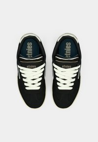 Μαύρα αθλητικά παπούτσια με λευκές γλώσσες, ακατέργαστο ύφασμα επάνω, επενδεδυμένος γιακάς και καουτσούκ σόλα. Λογότυπο Etnies τυπωμένο στην γλώσσα.