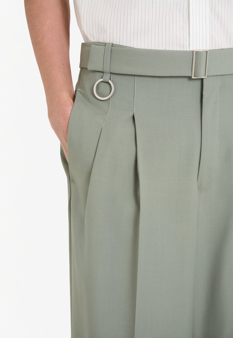 Pantalons plissés en tissu vert clair avec une ceinture ornée d'un détail en forme de boucle en argent. Texture lisse et unie avec des poches latérales.