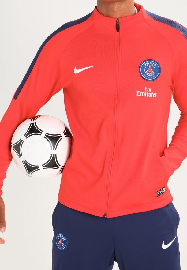 Punainen vetoketjullinen takki, jossa on tummansinisiä yksityiskohtia ja pyöreä Paris Saint-Germain -logo, yhdistetty tummansinisiin housuihin ja musta-valkoiseen jalkapalloon.