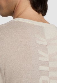 Maglione a maglia beige chiaro con colletto a coste. Presenta un motivo grafico sottile sul retro in una tonalità leggermente più scura.