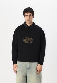 Suéter negro de pullover con cuello de cuarto de cremallera, que presenta un logo bordado en dorado en el frente y tela de cuello en gris contrastante.
