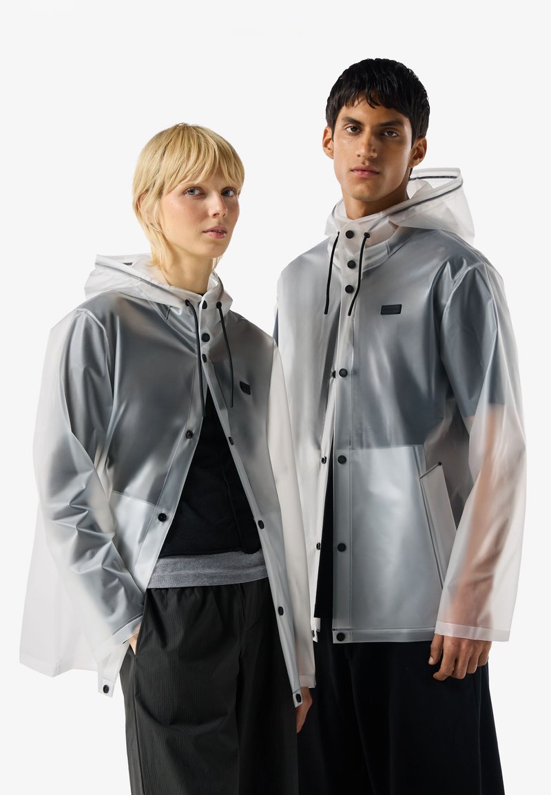 Zwei Models tragen durchsichtige graue Regenjacken mit schwarzen Knöpfen und Kapuzen mit Kordelzug, die nebeneinander vor weißem Hintergrund stehen.