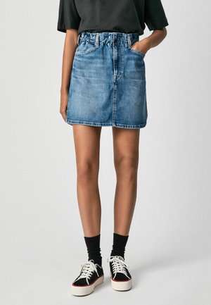 Frau trägt einen hoch taillierten blauen Jeansrock mit elastischem Bund, schwarze Socken, schwarz-weiße Sneakers und ein schwarzes kurzärmliges Shirt.