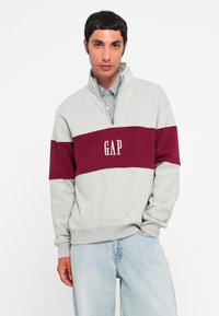 Gray sweatshirt featuring a maroon horizontal stripe, half-zip collar, and "GAP" logo in white. Made of soft fabric with relaxed fit.