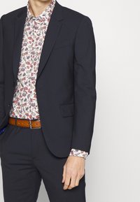 Chaqueta de traje azul marino sobre una camisa blanca con flores; la camisa presenta patrones rojos y azules; un cinturón de cuero marrón aporta contraste y detalle.