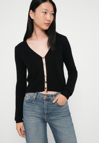 Jovem mulher a usar um cardigan preto com fechos em letras douradas a soletrar "FUTURO" e jeans azuis, a estar de pé com a mão no bolso.
