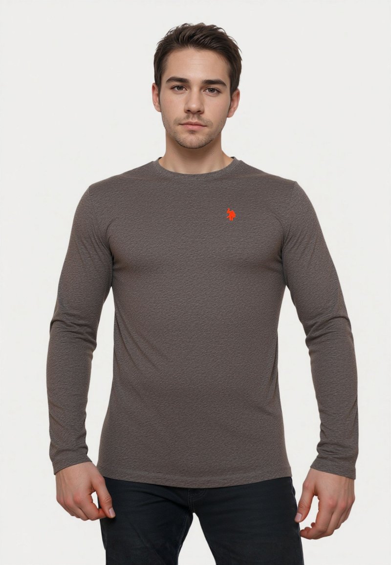 Young man with short dark hair wearing a fitted long-sleeve textured gray shirt with a small red logo on the chest.