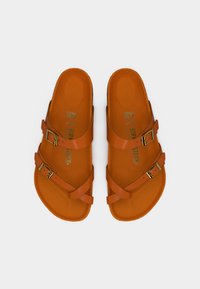 Ζευγάρι πορτοκαλί σανδάλια Birkenstock με δύο ρυθμιζόμενους ιμάντες και ορειχάλκινες αγκράφες, επίπεδη σόλα, εμφανίζονται από πάνω σε λευκό φόντο.